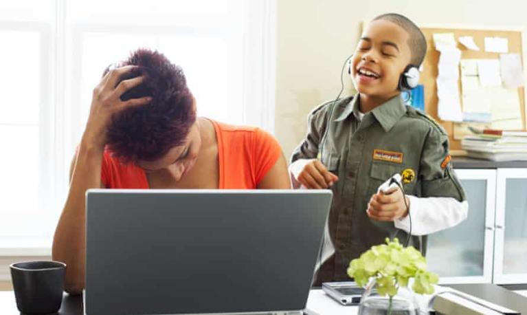 A mother at her laptop looking overwhelmed with her son dancing behind her. © Design Pics/Alamy